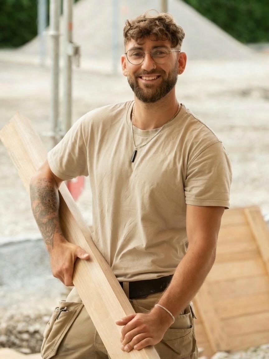 David Witt, Zimmerermeister, lächelnd auf der Baustelle mit einem Holzbrett im Arm